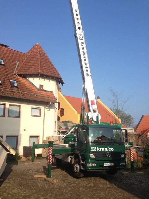 Bild ein grüner Autokran ist im Einsatz. Im Hintergrund sind 2 Gebäude.